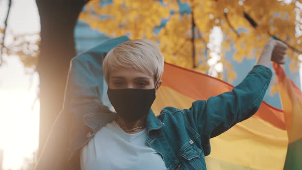 Young Blond Caucasian Woman with Face Mask Waving Rainbow Flag Under the Tree with Yellow Leaves