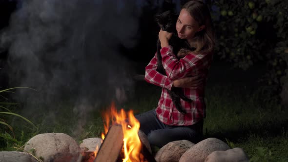 Woman Holds a Cat and Sits Near Campfire at Evening