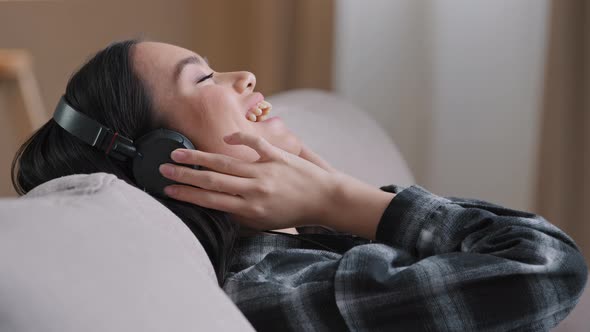 Calm Asian Korean Millennial Woman in Headphones Sitting Lying on Comfortable Home Sofa Couch with