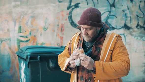 A Poor Man is Eating a Hamburger Next to the Waste Bins