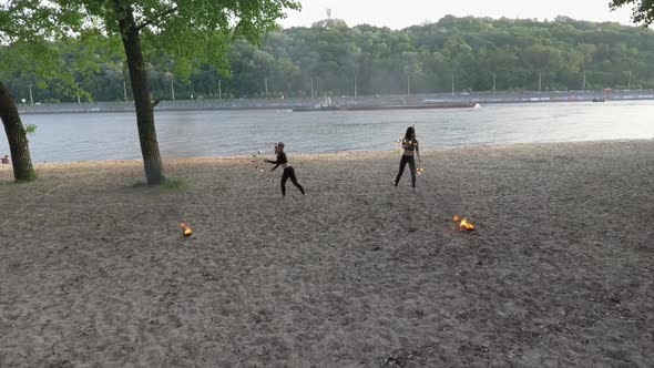 Two Young Women Performing a Show on the Sand with Flame Standing on the Riverbank