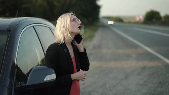 Confused Blonde in Glasses Talks on a Smartphone Emotionally at a Broken Car