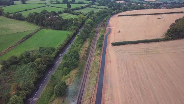 Wide aerial tracking backward along a train track and a road revealing a steam train just about to s