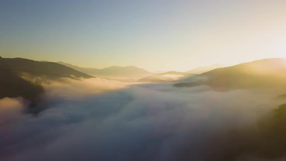 Aerial view of vibrant sunrise over white dense fog with distant dark silhouettes of mountain