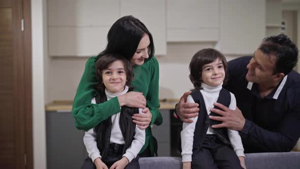 Smiling Parents Walking to Absorbed Twin Brothers Watching TV Sitting on Couch and Hugging Adorable