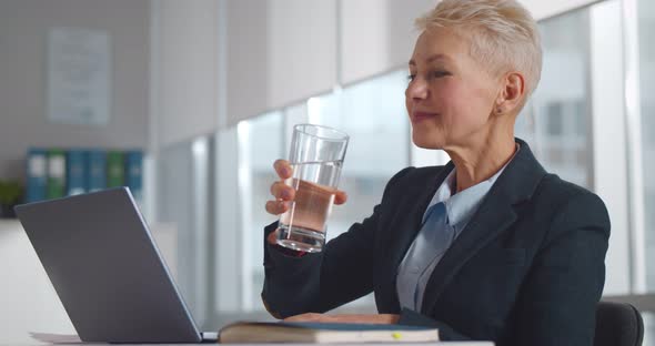 Senior Businesswoman Drinking Water Working on Laptop at Workplace