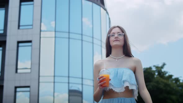 a Happy Girl in a Blue Dress and Glasses Drinks a Cocktail on the Street Against the Backdrop of a