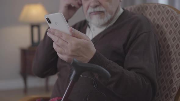 Unrecognizable Old Man Turning on Playlist on Smartphone and Listening To Music in Earphones