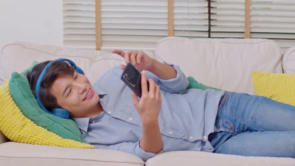 Asian handsome man lying on couch wearing headphone listen music and using mobile or smart phone