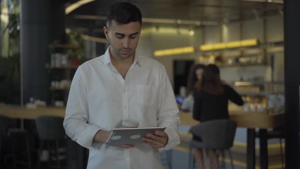 Portrait of Handsome Young Middle Eastern Man Using Tablet, Drinking Coffee, and Smiling at Camera
