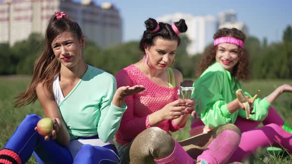 Retro Friends Teasing Young Woman Eating Chocolate Bar Outdoors Sitting on Sunny Lawn