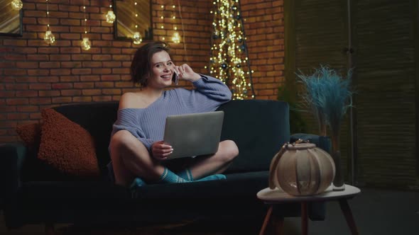 Girl Relaxing on a Sofa in Evening Holding Laptop and Speaking on Phone