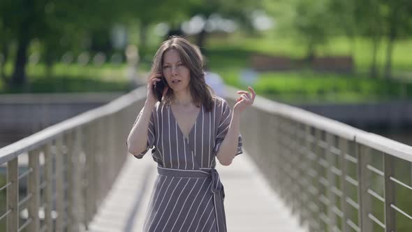 Middleaged Woman is Talking By Cell Phone Walking Over Small Bridge in Town at Summer Day