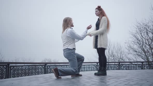 Side View of Stylish Boy in White Shirt and Vest Standing on One Knee and Proposing To His