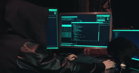 Hacker in a Hood Sits in Front of Computer Screens and Hacks Databases