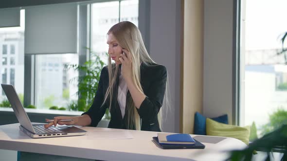 Trendy Business Woman Watching Laptop and Speaking on Phone. Stylish Young Blond Woman Working in