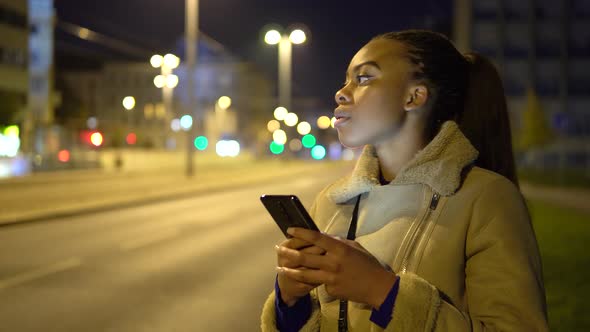 A Young Black Woman Works on a Smartphone and Then Smiles in a Street in an Urban Area at Night