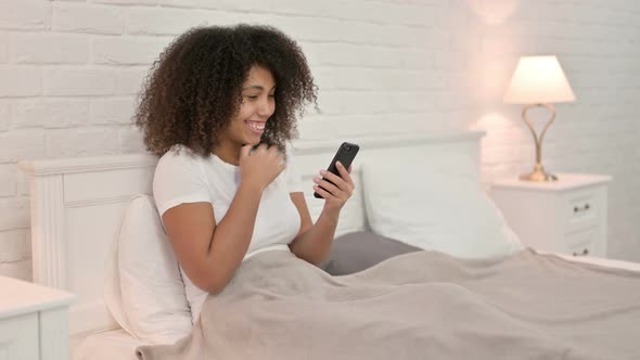 Success, Young African Woman Celebrating on Smartphone in Bed