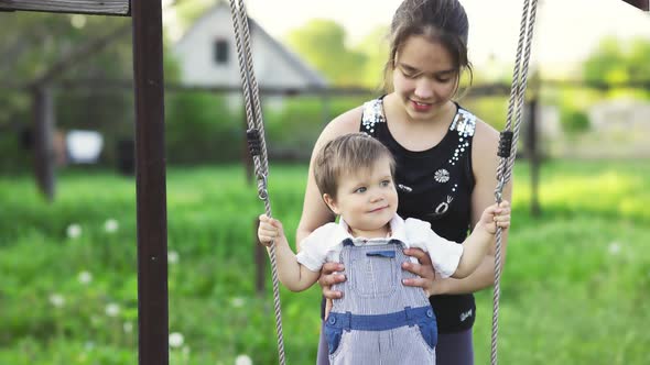 Cute Brother and Older Sister Ride on a Bright Swing on Warm Spring Day Against the Backdrop of a