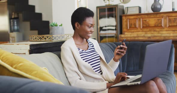 Happy african american woman sitting on sofa in living room, using laptop and smartphone