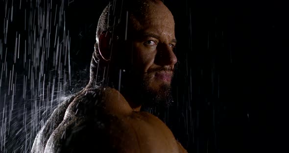 Close-up Portrait of a Brutal Bodybuilder in the Dark in the Rain Stands and Looks at the Camera