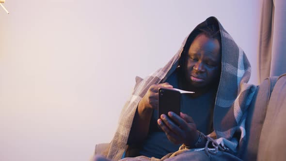 Ill African American Black Man Covered in Blanket Checking Temperature and Using Phone