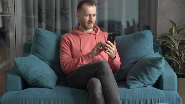 A Young European Man with a Smartphone on the Sofa in His Home Interior Uses Internet Messengers and