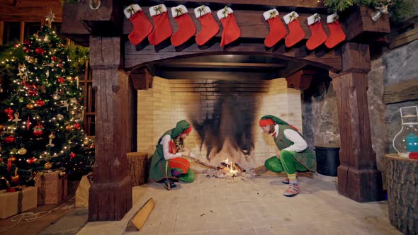 Elves near fireplace on Christmas background indoors. Funny elves in green costumes sit