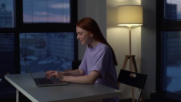 Cheerful Redhead Young Woman Freelancer Using Laptop Computer for Work Sitting at Desk Near Window