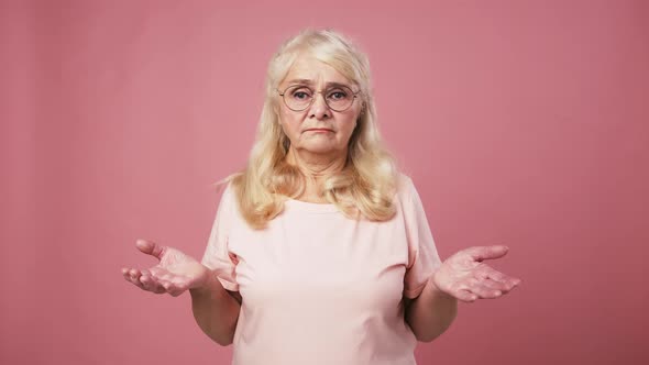 Puzzled Senior Lady in Glasses Shrugging Her Shoulders Gesturing Hands As Ignorance Pink Background