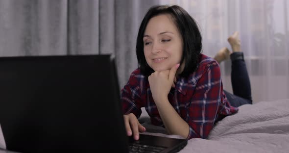 Young Brunette Woman Lying on the Bed. Chatting, Working on the Laptop Computer