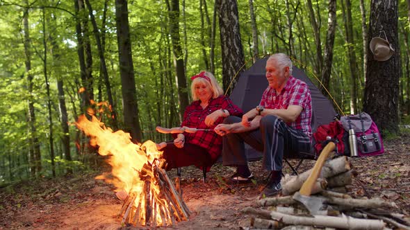 Senior Elderly Grandmother Grandfather Cooking Frying Sausages Over Campfire in Wood at Camping