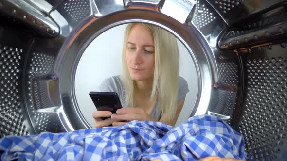 a woman looks at her smartphone through open door of washing machine.