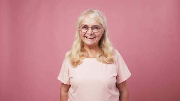 Positive Senior Woman in Glasses Gesturing Thumbs Up Shaking Hands and Smiling Pink Studio