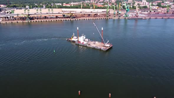 Tugboat pushing barge with view of industrial cranes of Klaipeda harbor, aerial view