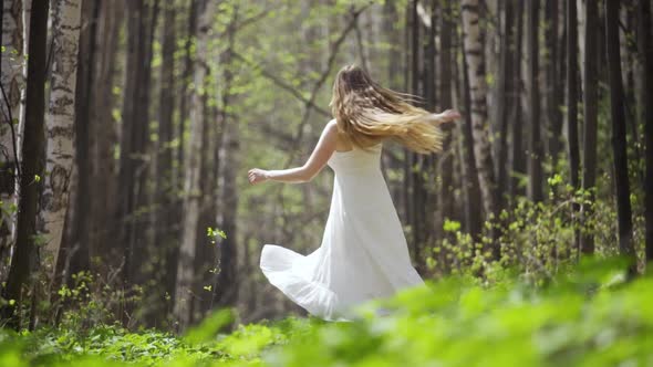 A Cute Young Woman Whirls Around Herself in a White Dress in a Light Forest