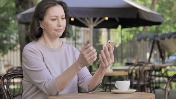 Old Woman Upset by Loss on Smartphone, Sitting in Outdoor Cafe