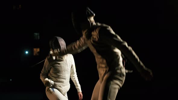 Two Fencers In Fencing Suits And Helmets Fencing In The Dark Outside.