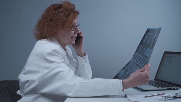 Elderly Caucasian Woman Doctor Analyze Xray Image and Talk with Mobile Phone About Diagnostics in