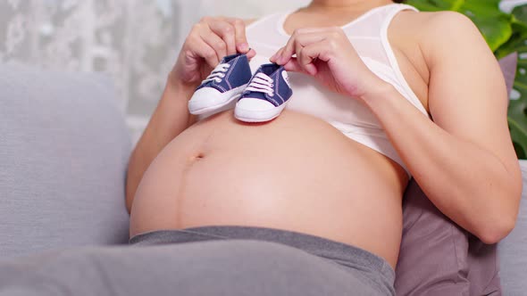 Happy Pregnant Woman lying on couch holding baby sneakers walking on her big belly at cozy home