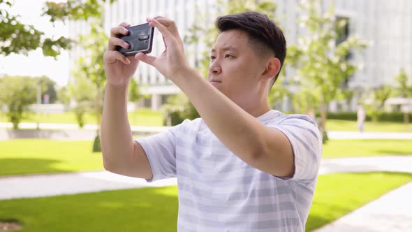 A Young Asian Man Takes Pictures with a Smartphone in an Urban Area  a Park and Office Buildings