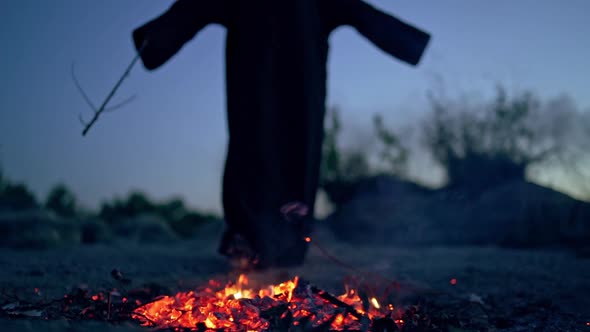 Spooky death in black gown near fire. Embers of fire on the ground and mysterious figure doing shama