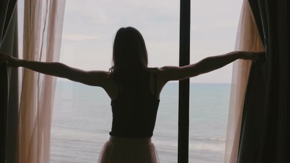 Young Happy Woman Opening Window Curtains in Dark Room To Enjoy Incredible Sea Waves View, Touching