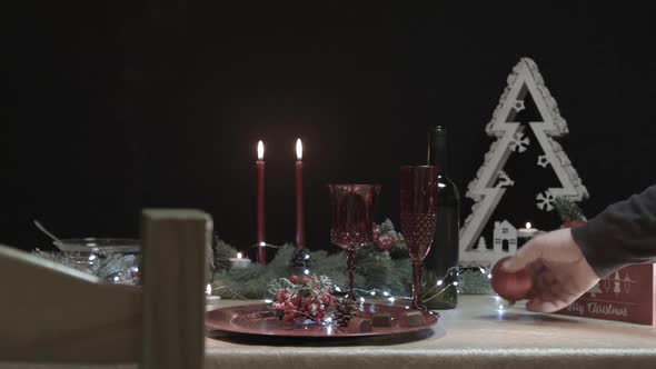 Man takes a red Christmas tree ball on a decorated table in a dark room with a chair in foreground