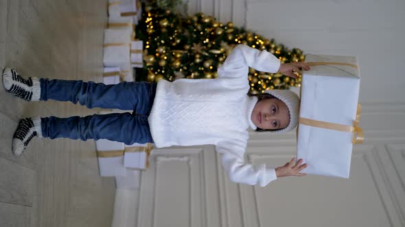 Child Boy in a White Knitted Sweater and Hat Holds Gift Box with a Gold Ribbon on His Head