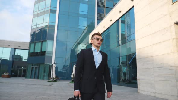 Young Businessman in a Black Suit with Briefcase Walking Along Modern Office Building. Confident