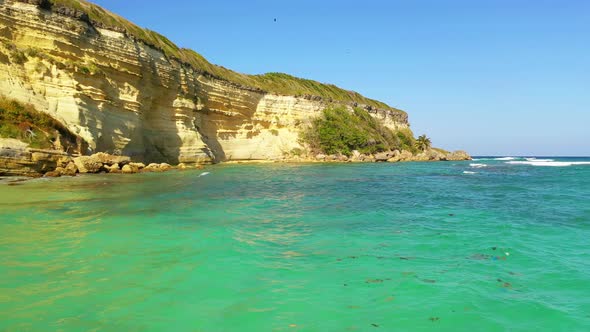 Stunning low flight over the rocky sandy beach towards the waves of the turquoise ocean and the stee