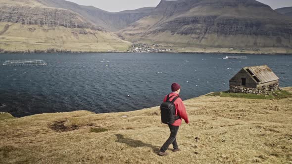 Man Walking Towards Wooden Shed By Cliff in Faroe Islands - Sea in Background