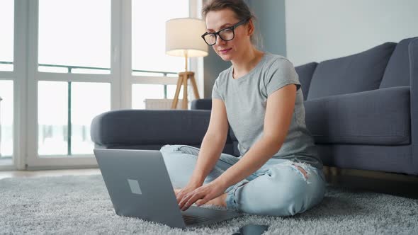 Casually Dressed Woman Sitting on Carpet with Laptop and Smartphone and Working in Cozy Room