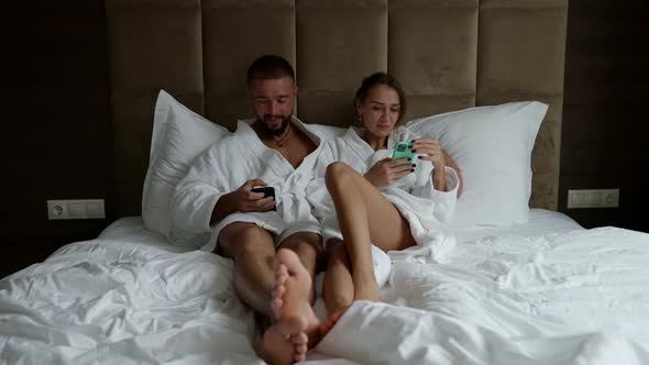 a Couple in White Coats is Lying in Bed with Phones